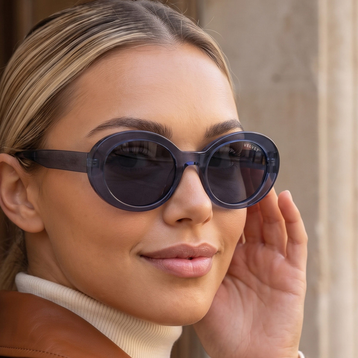 Model wearing Velvet Eyewear® Iris polarized sunglasses in navy blue, a bold round oval frame in translucent navy acetate with smoke grey polarized lenses and the VELVET wordmark on the temple, photographed against a sunlit limestone wall. Frame shape: Round Oval. Suits oval, square and heart face shapes. Colour palette: Winter, Summer, Autumn, Spring. Navy Blue Polarized. Lifestyle image.