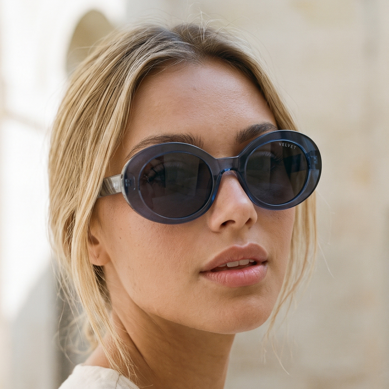 Model wearing Velvet Eyewear® Iris polarized sunglasses in navy blue, a bold round oval frame in translucent navy acetate with smoke grey polarized lenses and the VELVET wordmark on the temple, photographed against a sunlit limestone wall. Frame shape: Round Oval. Suits oval, square and heart face shapes. Colour palette: Winter, Summer, Autumn, Spring. Navy Blue Polarized. Lifestyle image.