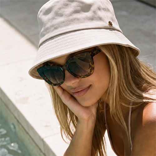 Velvet Trends UPF50 bucket hat in sand on our model Mia wearing the Velvet Eyewear sunglass style Maya in tortoise  while sitting on the edge of a pool.