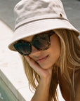 Velvet Trends UPF50 bucket hat in sand on our model Mia wearing the Velvet Eyewear sunglass style Maya in tortoise  while sitting on the edge of a pool.